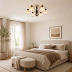Clérachic floral-glass chandelier over a bed with a rust and taupe throw and a round upholstered bench, with sheer curtains beside.