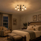 Warm-lit Clérachic chandelier in an evening bedroom with a wooden bed, a mustard armchair and framed botanical prints.