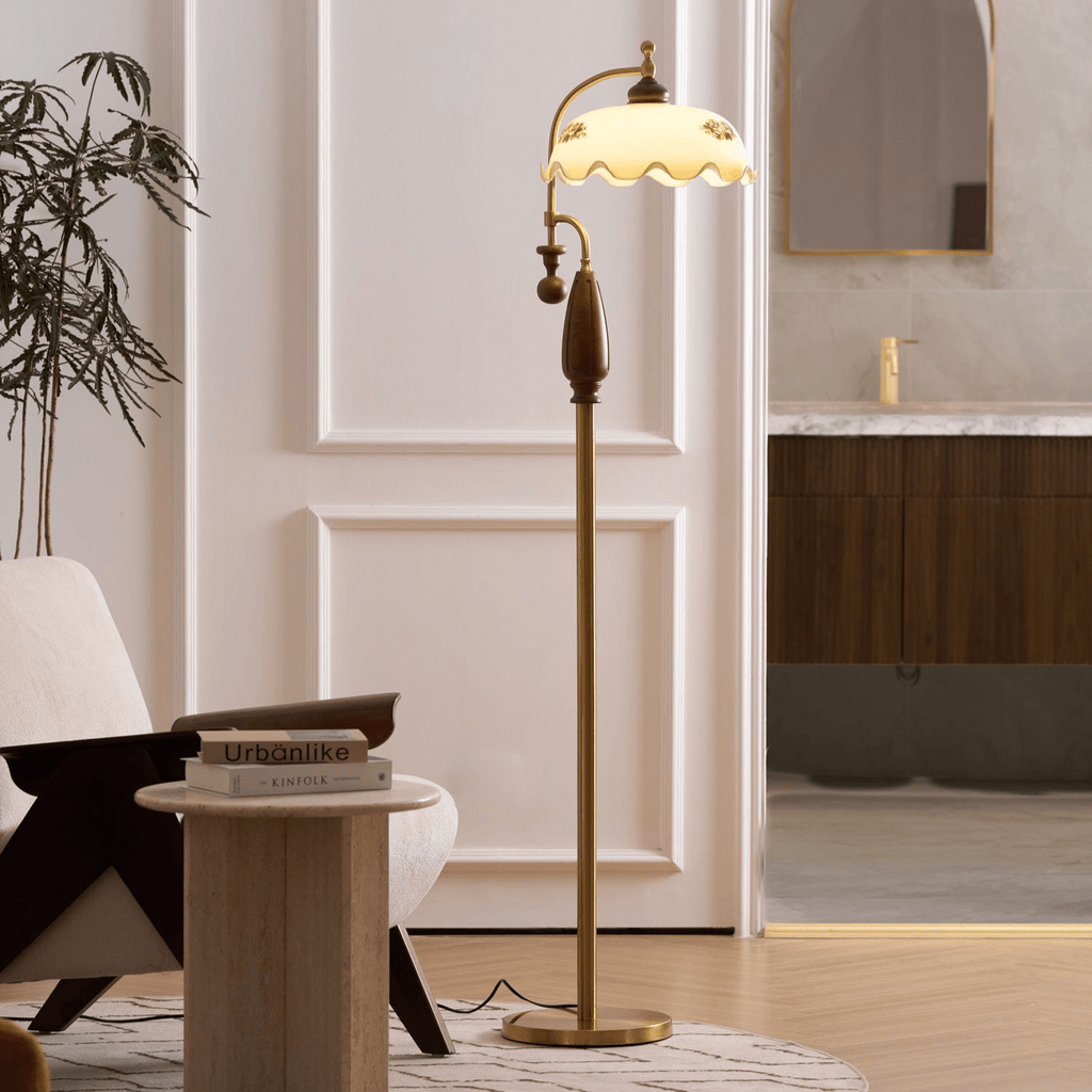 A slim brass floor lamp glows beside a white panelled door and a travertine side table; a Clérachic lamp with a frilled cream shade.