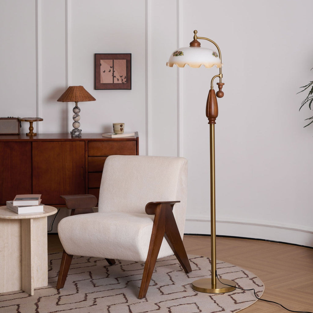Lamp seen from the left, its frilled glass shade glowing above a round brass base, near a boucle armchair on a patterned rug; a Clérachic floor lamp.