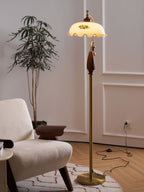 Brass stem and round base of this Clérachic floor lamp, with a black cord running across a patterned rug and a tall plant behind.