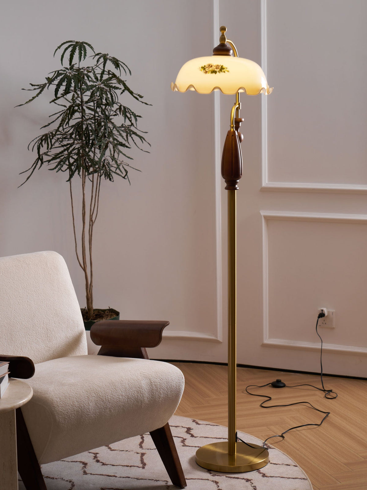 Brass stem and round base of this Clérachic floor lamp, with a black cord running across a patterned rug and a tall plant behind.