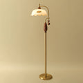 Brass and walnut version of the floor lamp on a warm gold backdrop, shade unlit, with its round brass base on the floor; a Clérachic lamp.