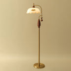 Brass and walnut version of the floor lamp on a warm gold backdrop, shade unlit, with its round brass base on the floor; a Clérachic lamp.