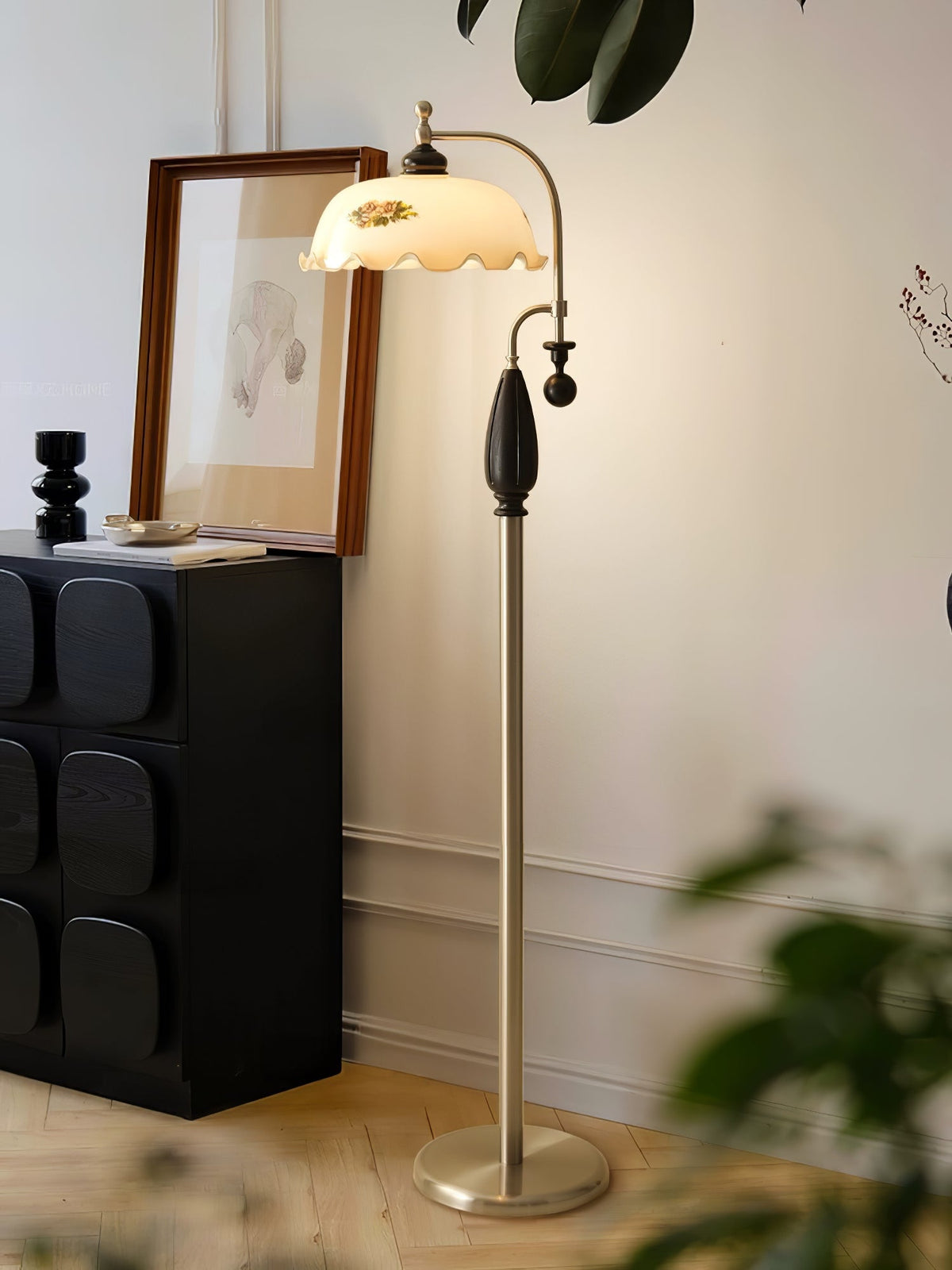 Black-and-nickel floor lamp lit beside a dark carved cabinet and a framed sketch, with leaves in the foreground; a Clérachic lamp.