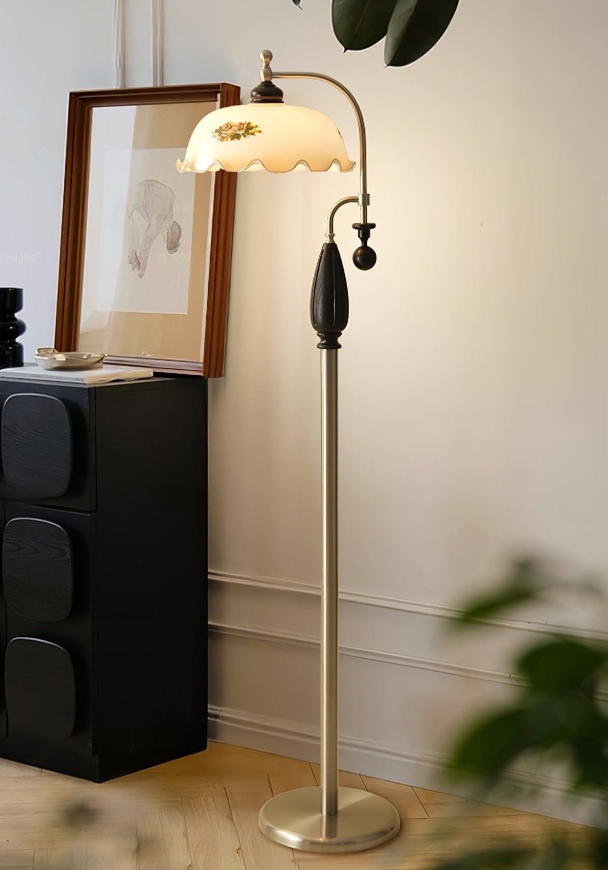 Nickel and black version of this Clérachic floor lamp lit beside a dark carved cabinet, a framed sketch and a leafy plant.