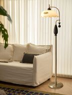 Nickel floor lamp with a black grip stands beside a linen sofa, in front of vertical white blinds; a Clérachic design.