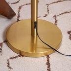 Close view of the round brass base and the black cord leaving the stem, on a patterned cream rug; a Clérachic floor lamp.