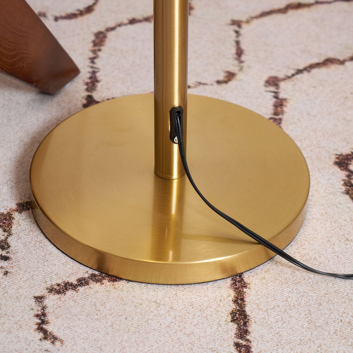 Close view of the round brass base and the black cord leaving the stem, on a patterned cream rug; a Clérachic floor lamp.