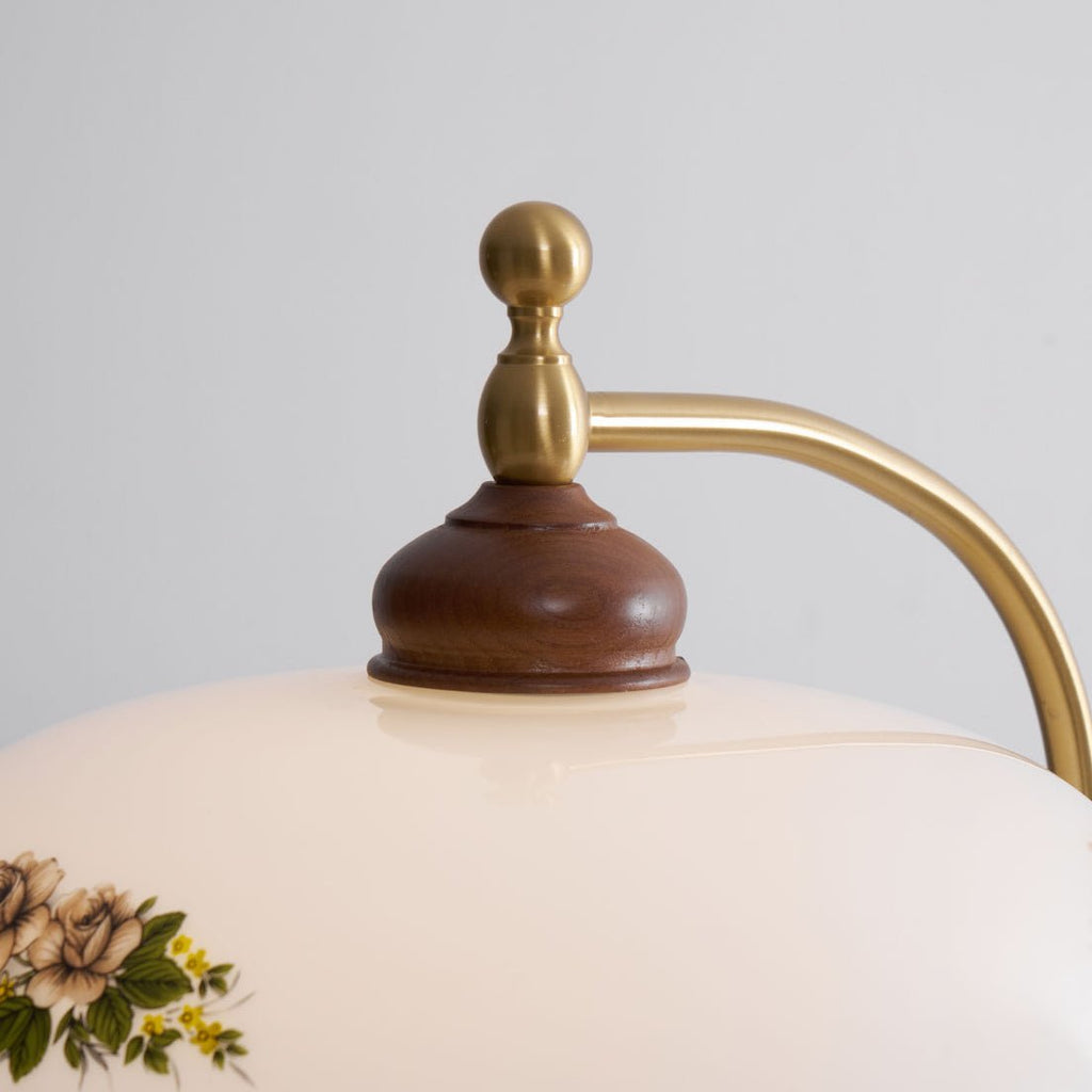 Close view of the brass finial, curved arm and walnut cap sitting on the cream glass, with floral print below; a Clérachic floor lamp.