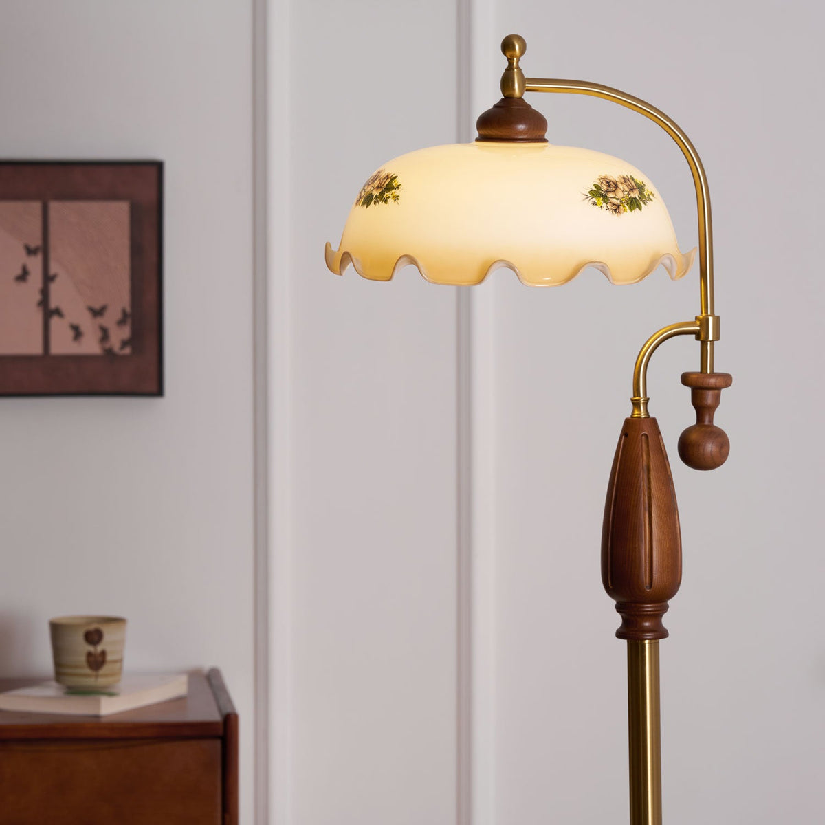 Lit shade with two floral prints, shown above the walnut grip and brass stem, with a framed print and a small cup behind; a Clérachic lamp.