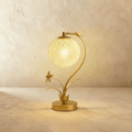 Clérachic gold table lamp with a curved stem and a lit woven sphere shade, a metal flower and leaves rising from a round base.