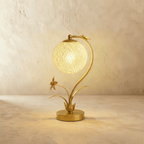 Clérachic gold table lamp with a curved stem and a lit woven sphere shade, a metal flower and leaves rising from a round base.