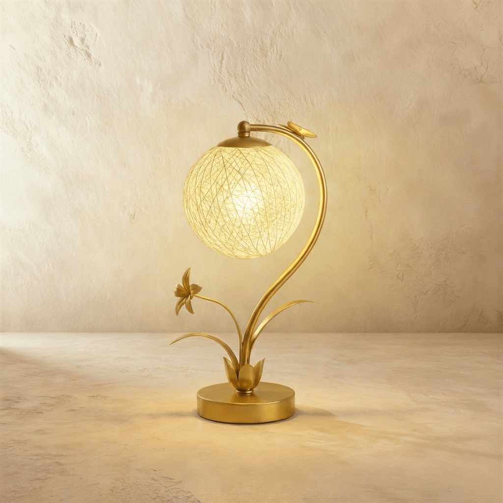 Clérachic gold table lamp with a curved stem and a lit woven sphere shade, a metal flower and leaves rising from a round base.