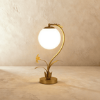 Clérachic gold table lamp with a glowing plain opal white sphere on a curved stem, metal flower and leaves at the base.
