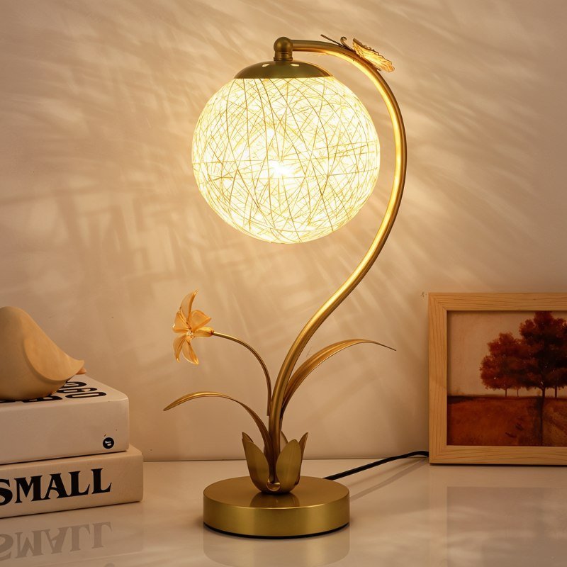 Clérachic gold table lamp with a lit woven sphere on a white desk beside books, a ceramic bird and a framed tree print.