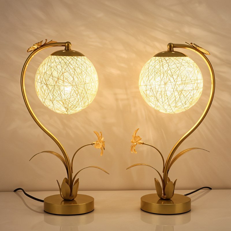 Pair of Clérachic gold table lamps with lit woven spheres and flower-and-leaf bases, on a white surface against a warm wall.