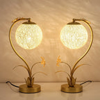 Pair of Clérachic gold table lamps with lit woven spheres and flower-and-leaf bases, on a white surface against a warm wall.