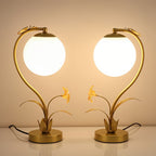 Pair of Clérachic gold table lamps with glowing plain white spheres and flower-and-leaf bases on a white surface.