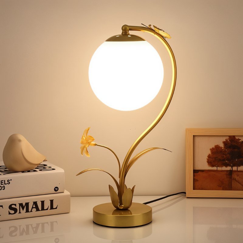 Clérachic gold table lamp with a lit plain white sphere on a white desk beside books, a ceramic bird and a framed tree print.