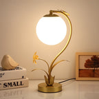 Clérachic gold table lamp with a lit plain white sphere on a white desk beside books, a ceramic bird and a framed tree print.