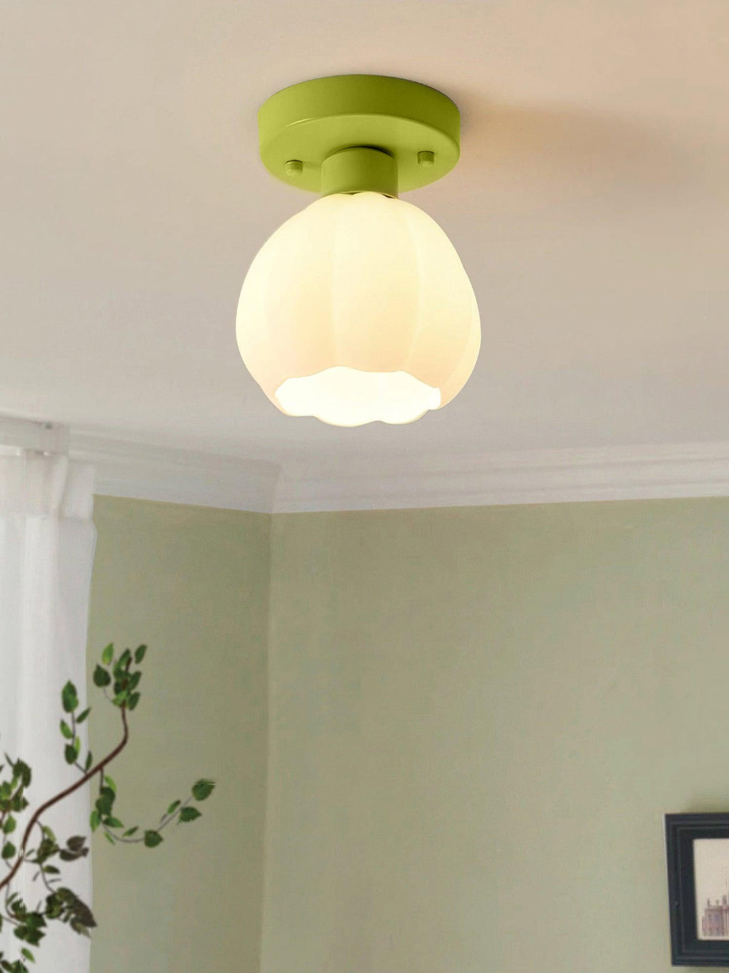 A lit Clérachic semi-flush light with a green canopy and a scalloped white glass shade, above a sage-green wall and a leafy plant.