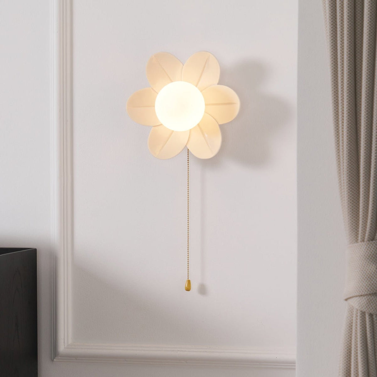 This Clérachic flower wall light glows softly on a panelled white wall, with a gold pull chain and a cream curtain at the right.