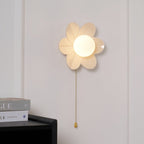 Lit flower-shaped wall light with a frosted globe centre, this Clérachic sconce hung above a black cabinet and stacked magazines.