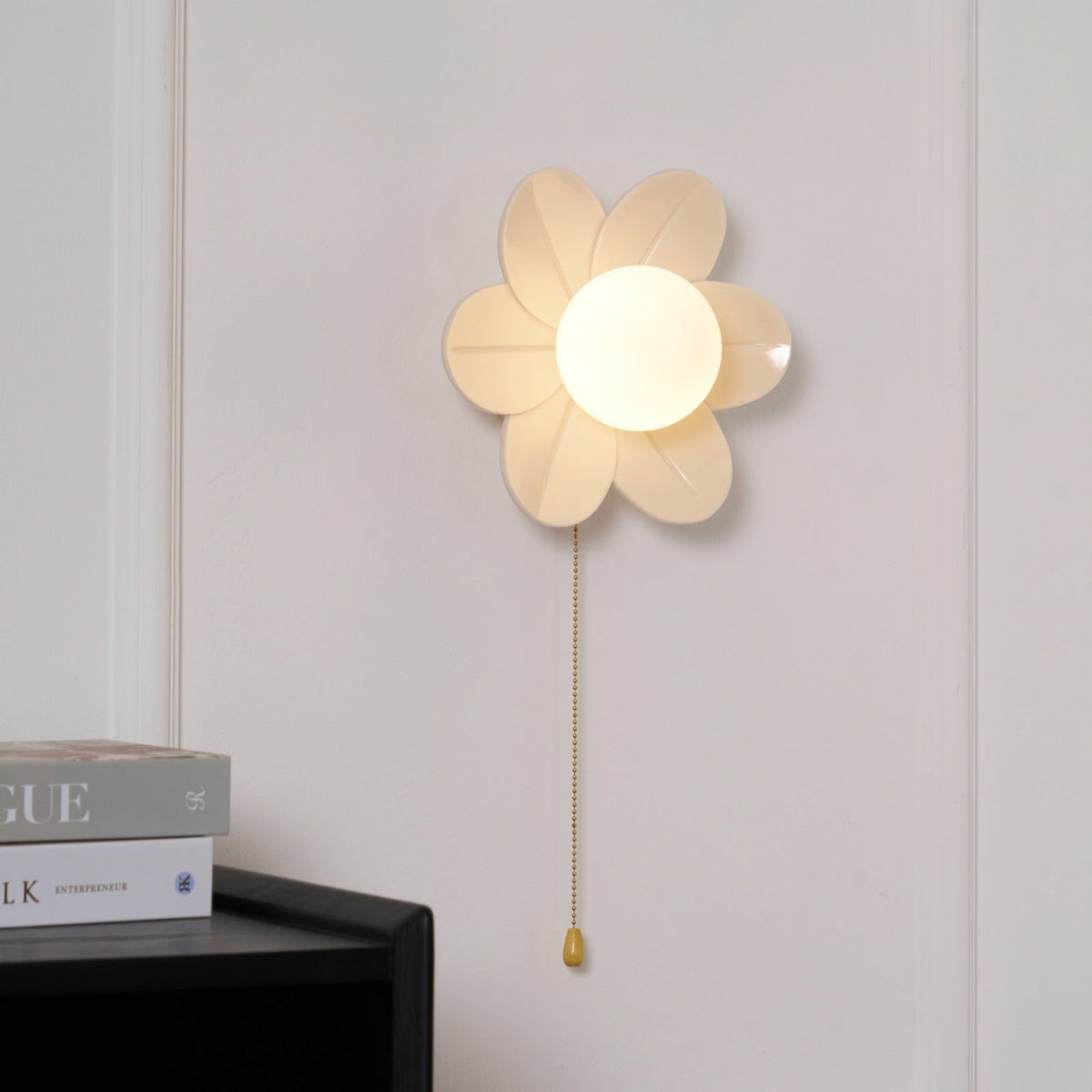 Lit flower-shaped wall light with a frosted globe centre, this Clérachic sconce hung above a black cabinet and stacked magazines.