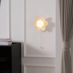 A small Clérachic flower wall light lit on a moulded white wall, beside a black cabinet and a taupe curtain.