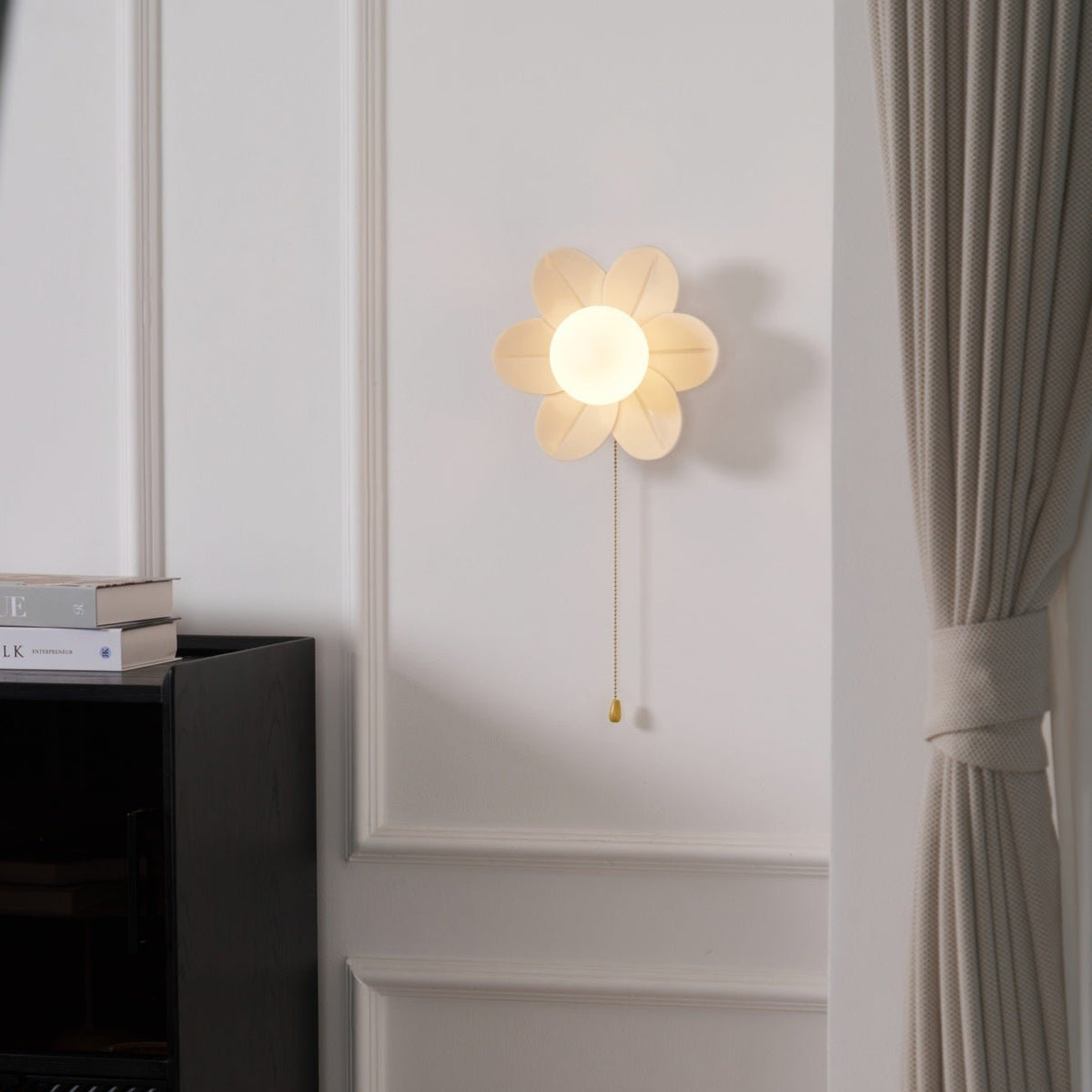 A small Clérachic flower wall light lit on a moulded white wall, beside a black cabinet and a taupe curtain.