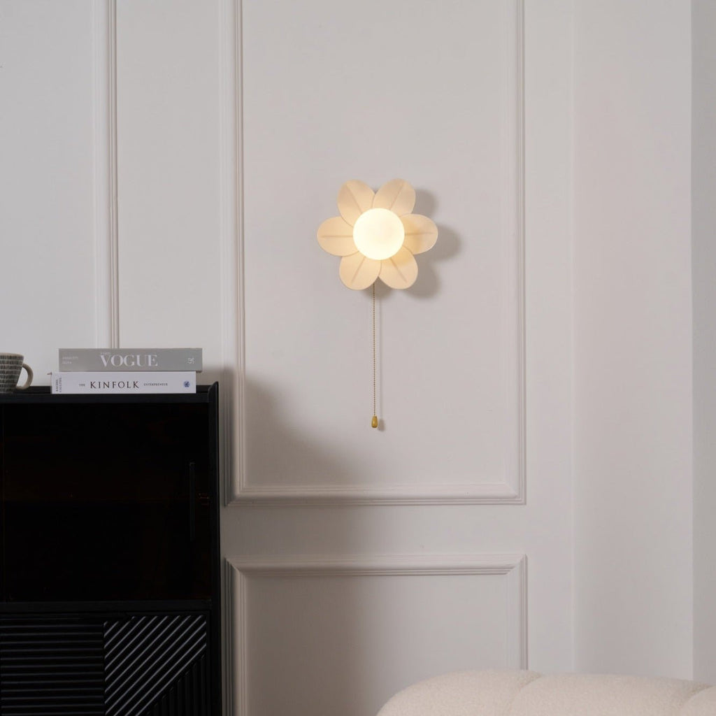 Flower wall light from Clérachic glowing gently on a white panelled wall, above a black cabinet and a pair of magazines.