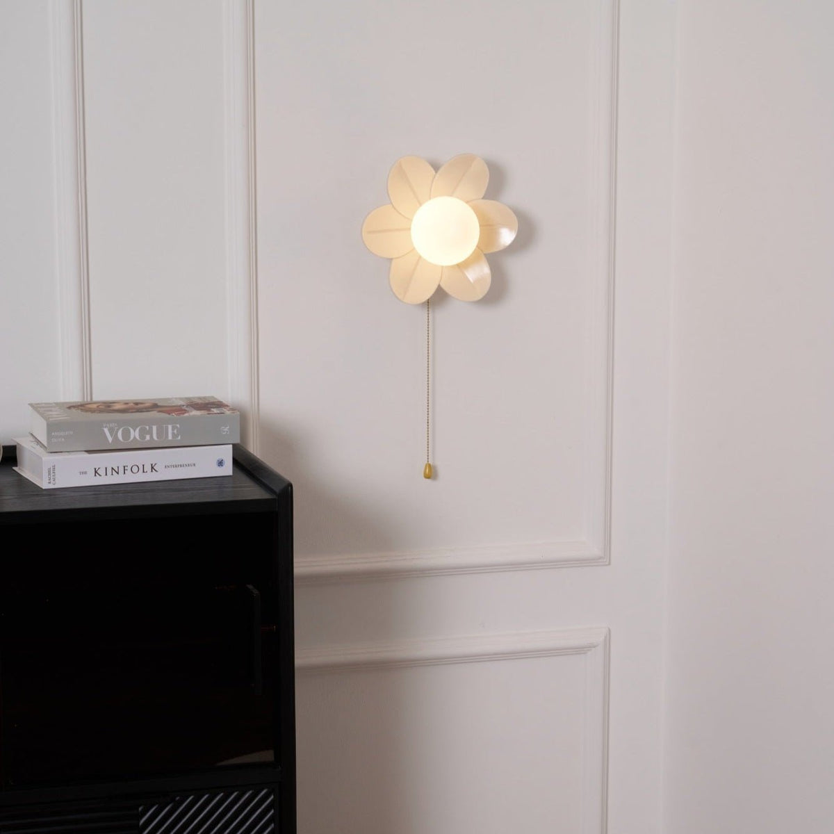 Small lit Clérachic flower sconce on a white wall panel, with a gold chain pull and a stack of magazines on the cabinet below.