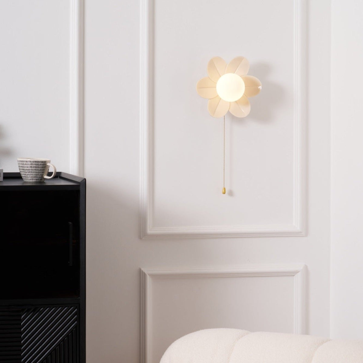 Lit petal sconce from Clérachic on a white panelled wall, with a coffee cup on the black cabinet and a cream sofa edge in view.