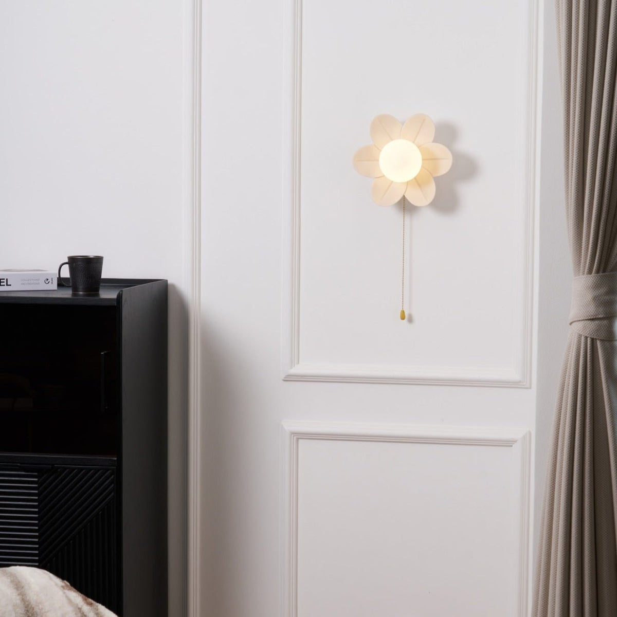 Clérachic flower wall light lit above a black cabinet with a mug on top, a heavy curtain hanging at the right edge of the wall.
