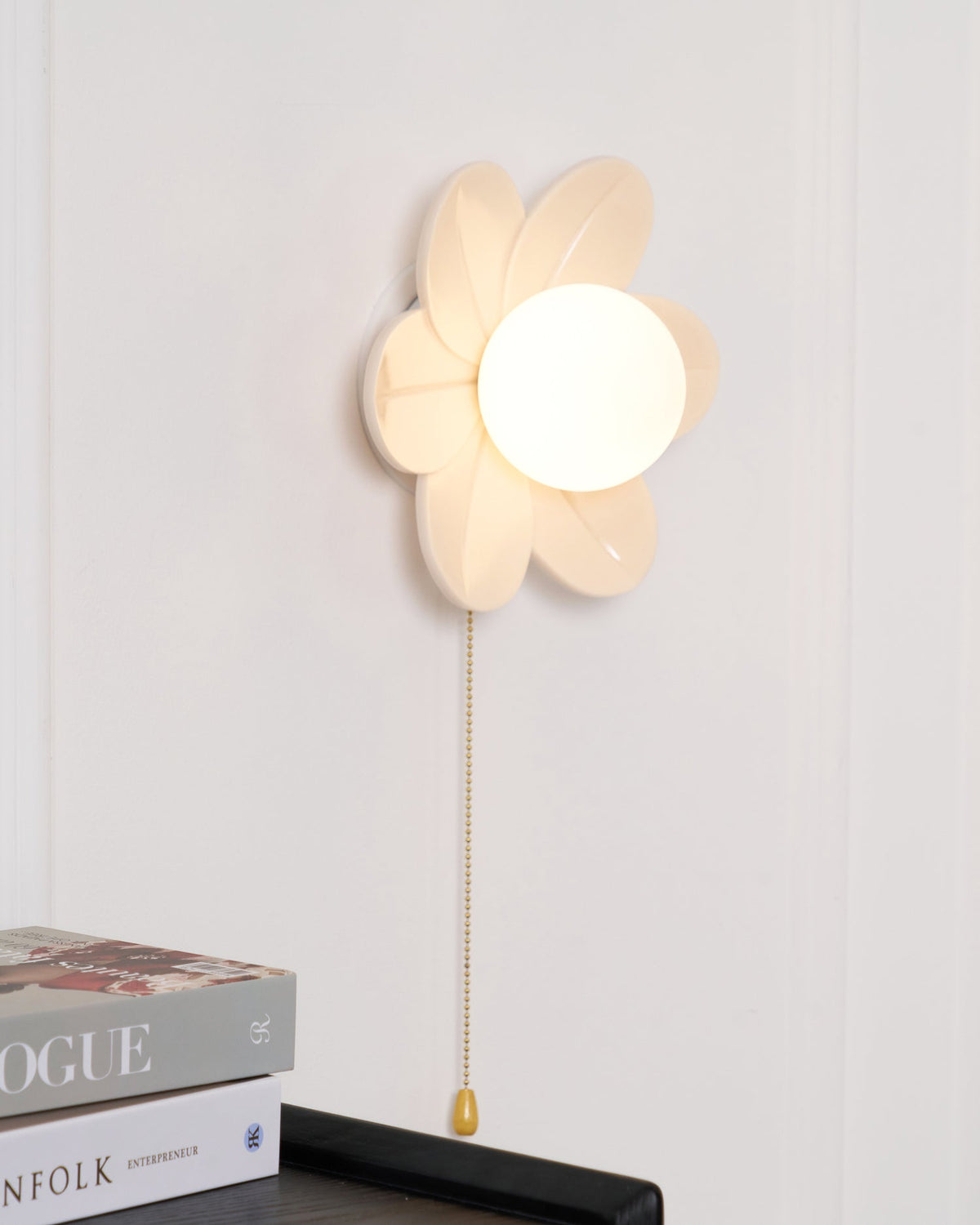 Side-angled view of the layered petals on this Clérachic flower wall light, with a gold chain falling past a black shelf and magazines.