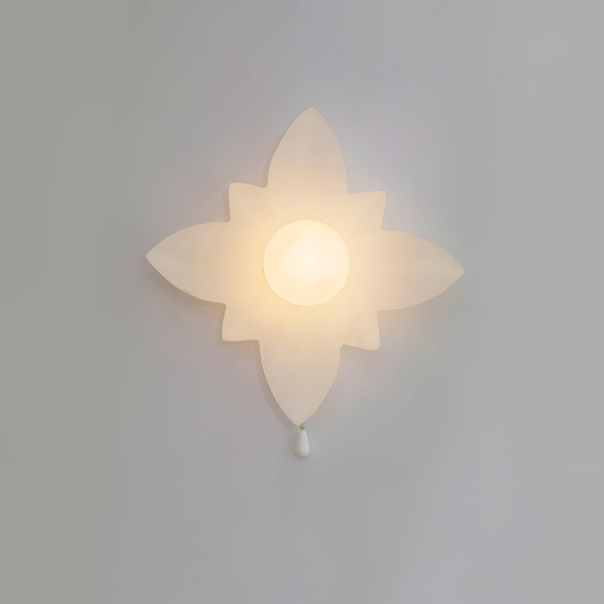 Four-point star-shaped Clérachic wall light lit warmly on a grey wall, with a frosted globe at the centre and a white pull below.