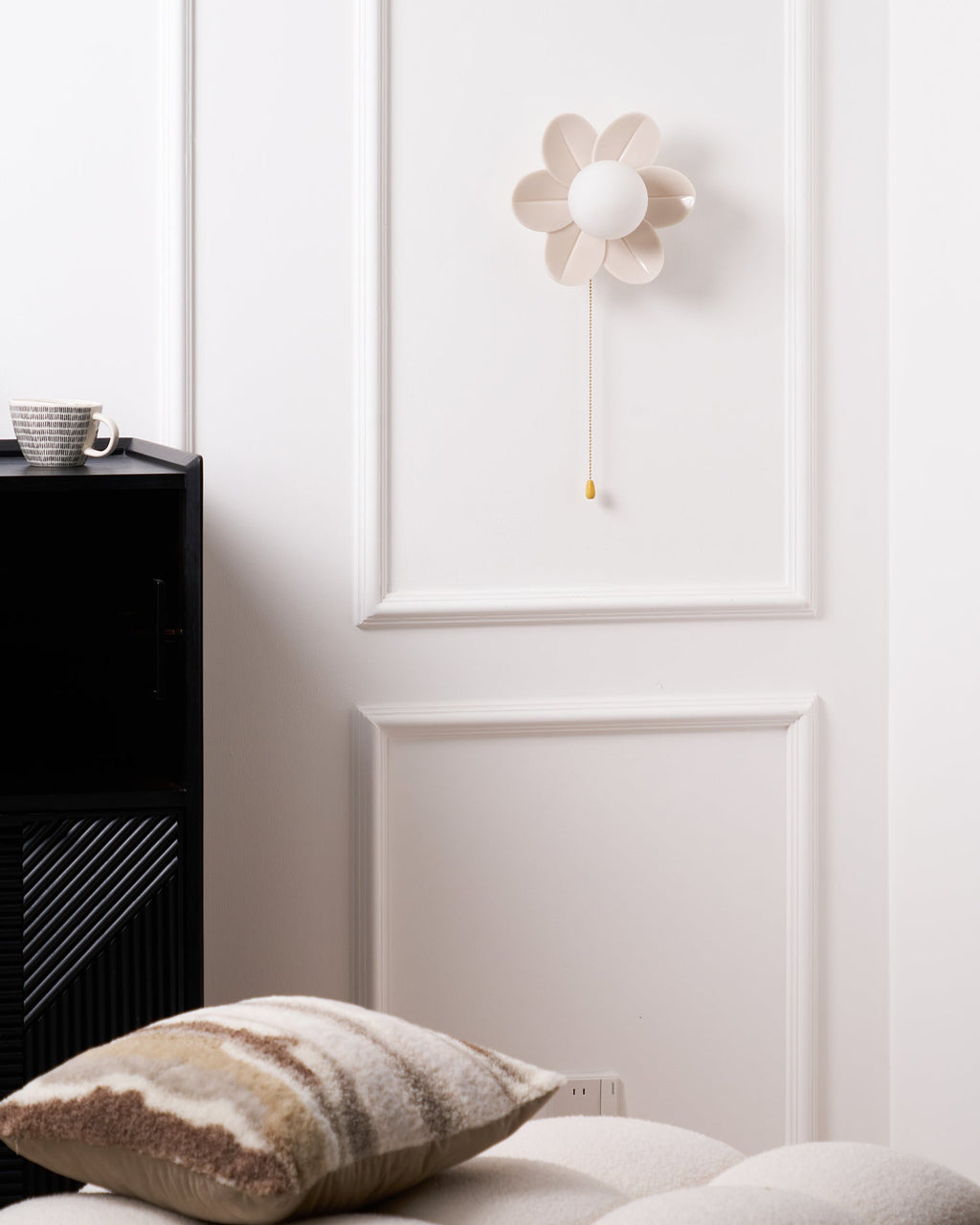 Small Clérachic flower wall light unlit on a panelled white wall, above a black cabinet with a mug and a cushion in view.