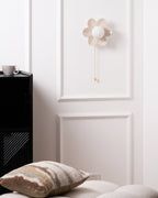 Small Clérachic flower wall light unlit on a panelled white wall, above a black cabinet with a mug and a cushion in view.