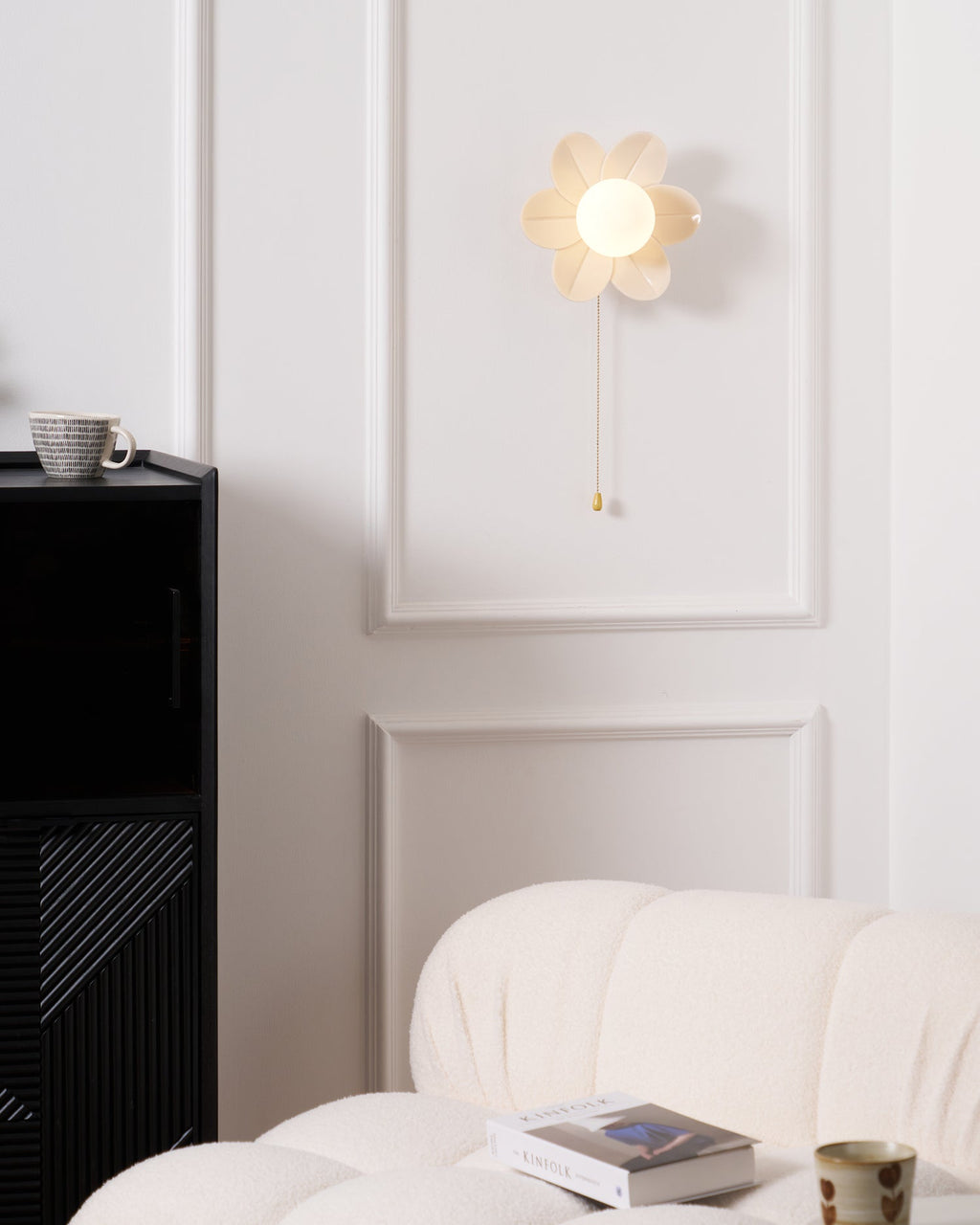 Lit flower sconce from Clérachic on a panelled white wall, above a black cabinet and a boucle sofa with a magazine and a mug.