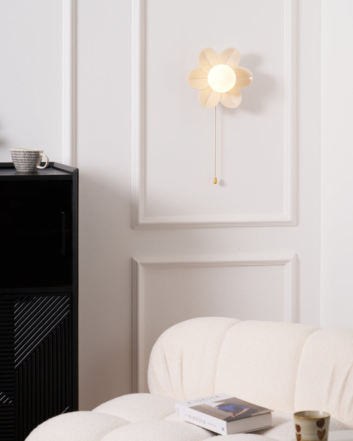 Lit flower sconce from Clérachic on a panelled white wall, above a black cabinet and a boucle sofa with a magazine and a mug.