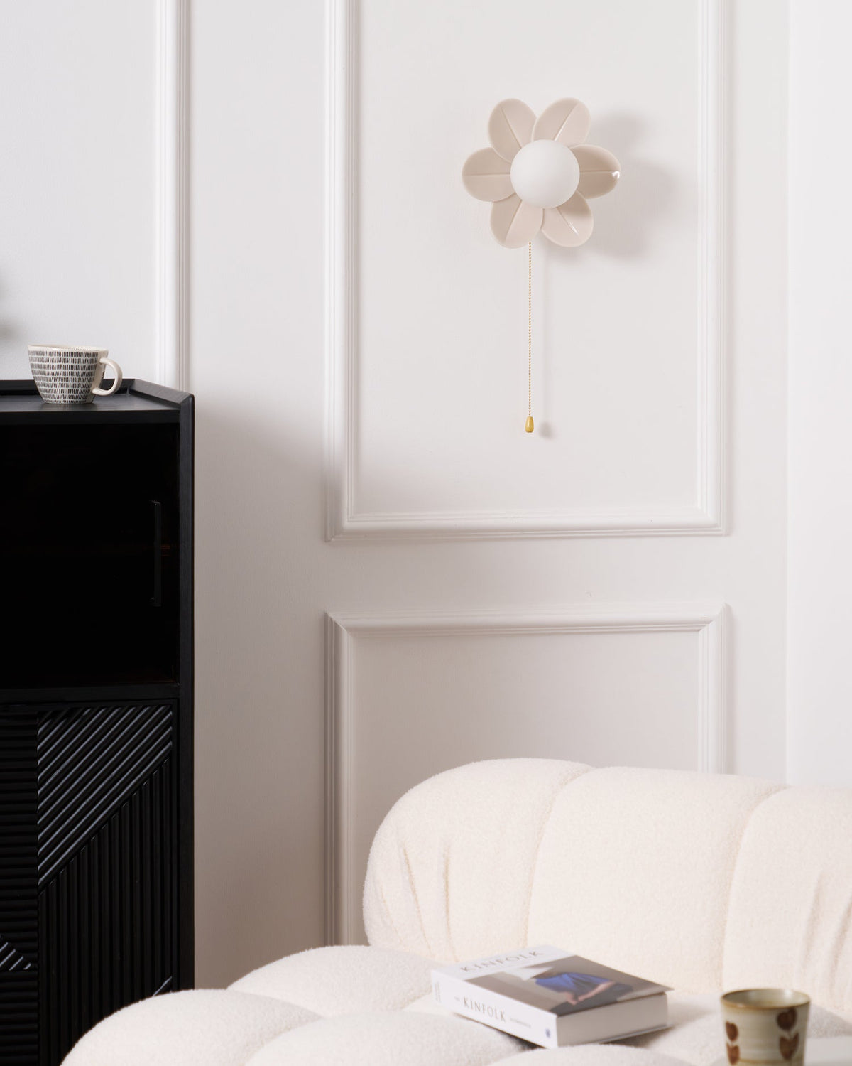 Clérachic flower wall light unlit on a white panelled wall, with a black cabinet and a mug at its side and a boucle sofa below.