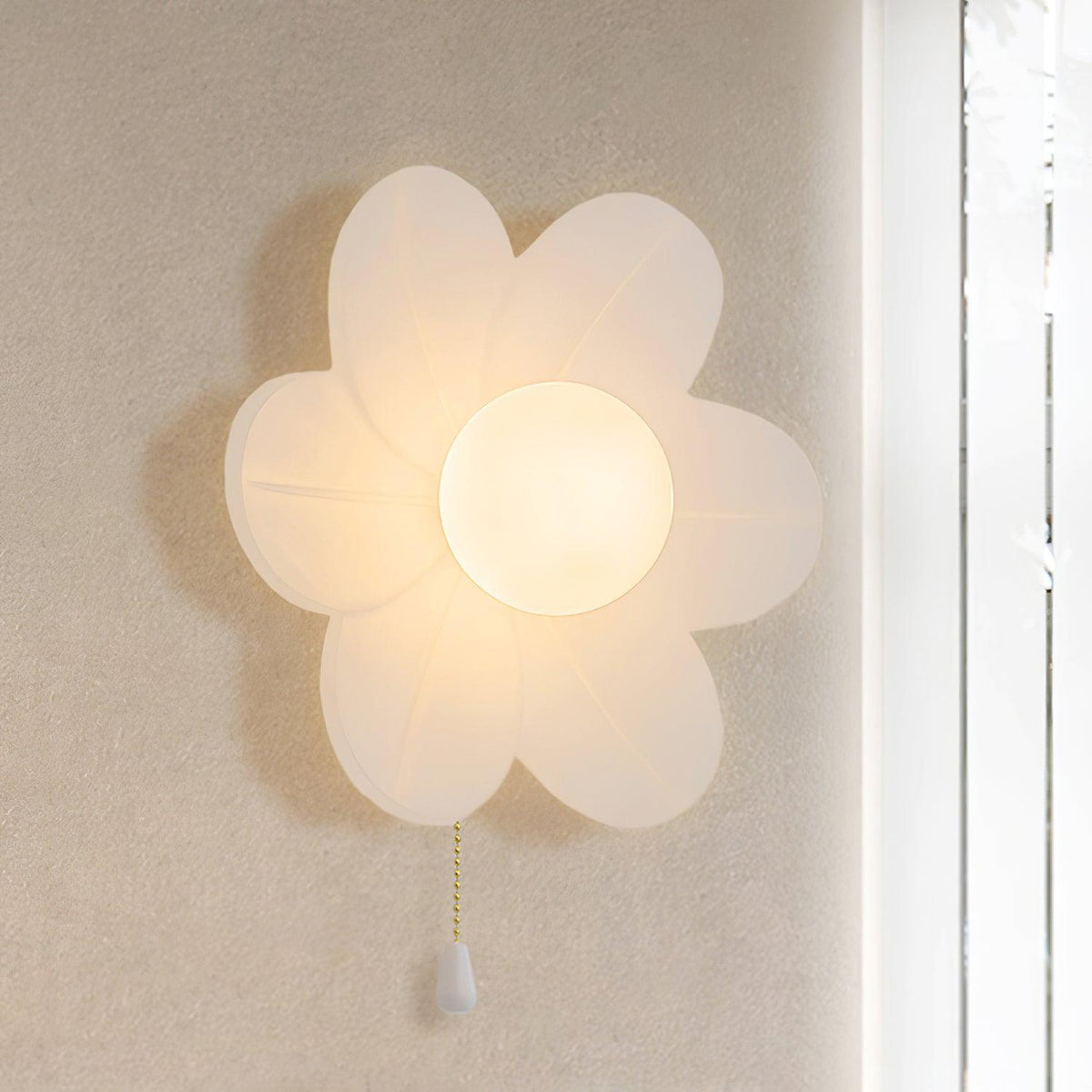 This Clérachic flower wall light glows warmly on a pale wall, its six petals translucent and a gold chain hanging down.