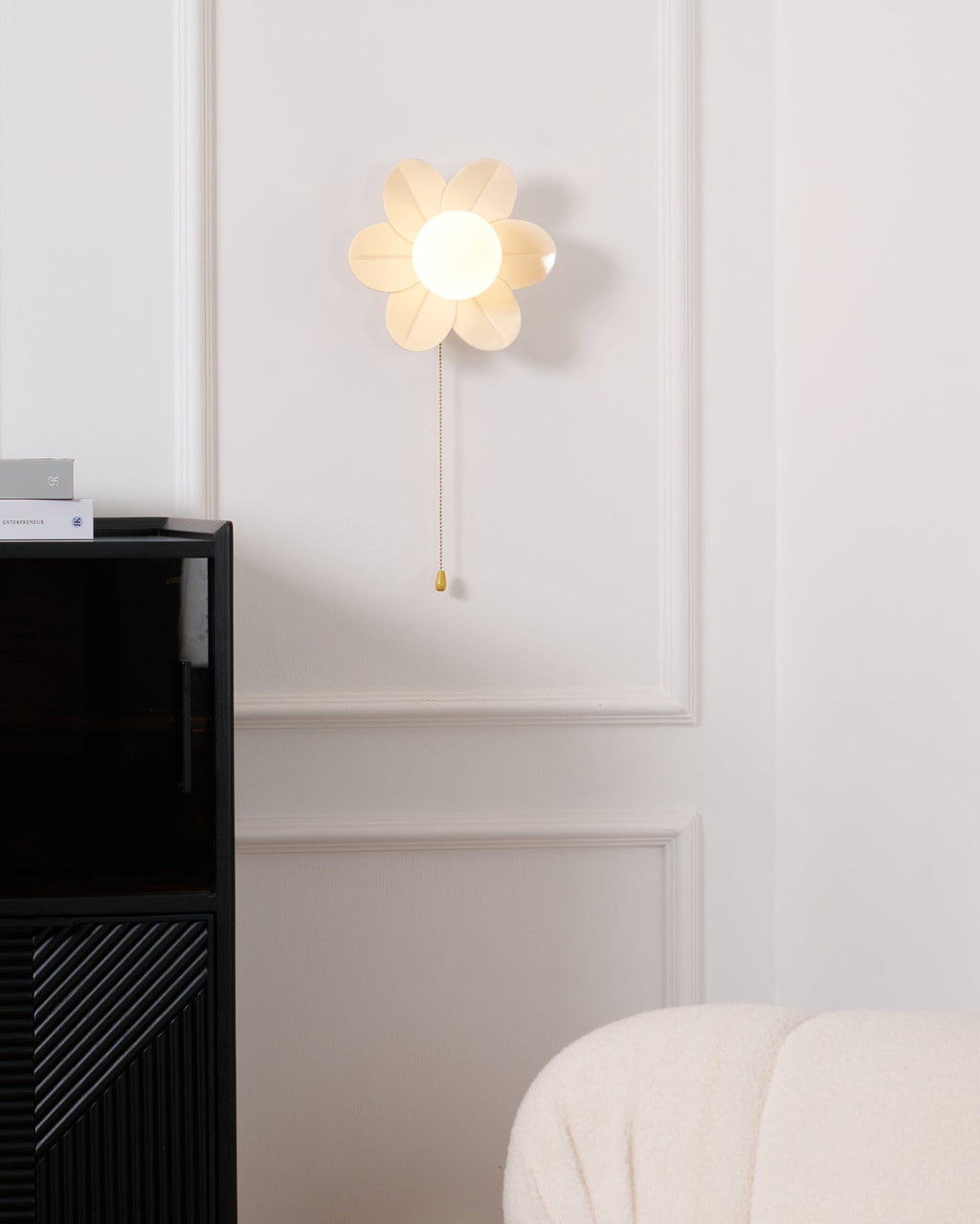 Clérachic flower wall light with a gold pull chain, hung above a black cabinet and a boucle sofa with a magazine and a mug.