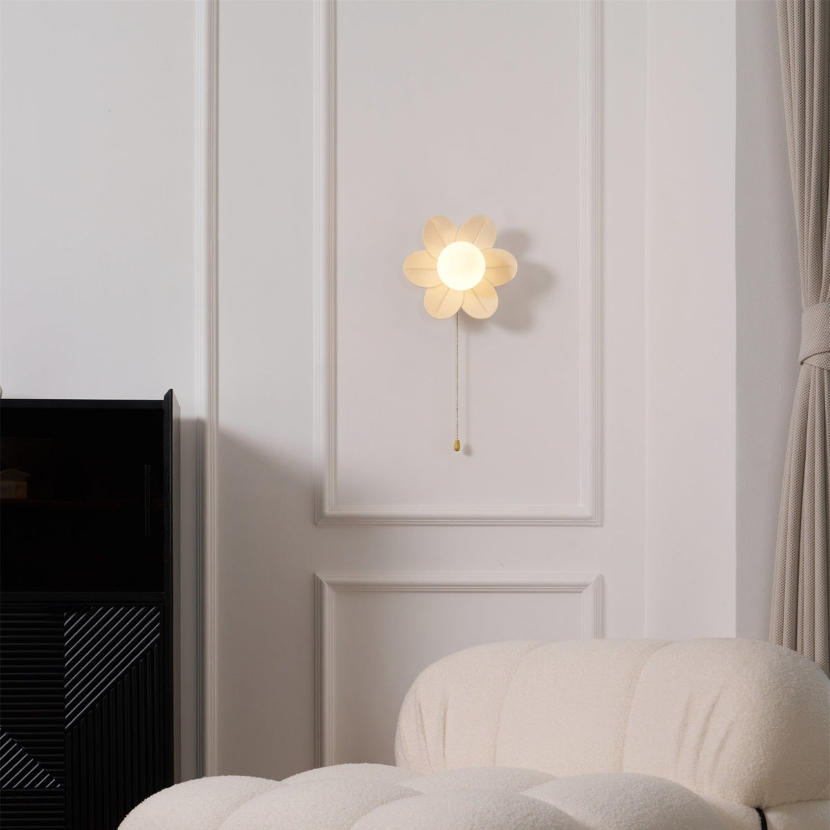 Flower wall light from Clérachic glowing on a moulded white wall, above a black cabinet and the arm of a boucle sofa.