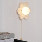 Warm-lit Clérachic flower wall light on a white wall, its petals tilted toward the viewer, above a Vogue magazine and a black cabinet.