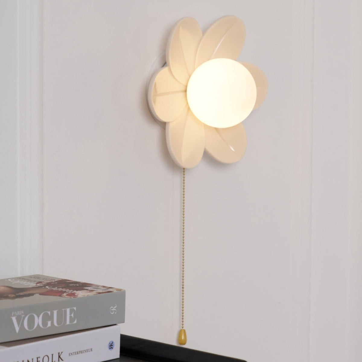 Warm-lit Clérachic flower wall light on a white wall, its petals tilted toward the viewer, above a Vogue magazine and a black cabinet.