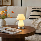 Lit on a round walnut coffee table beside an open magazine, this Clérachic mushroom lamp sits before a cream sofa and a vase of orange flowers.