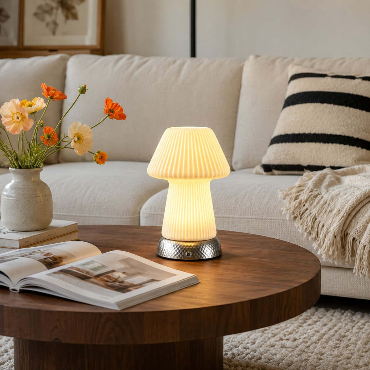 Lit on a round walnut coffee table beside an open magazine, this Clérachic mushroom lamp sits before a cream sofa and a vase of orange flowers.