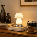 Lit on a pile of books on a wooden sideboard, this Clérachic lamp stands among dark glass vases, a vintage camera and a coffee cup.